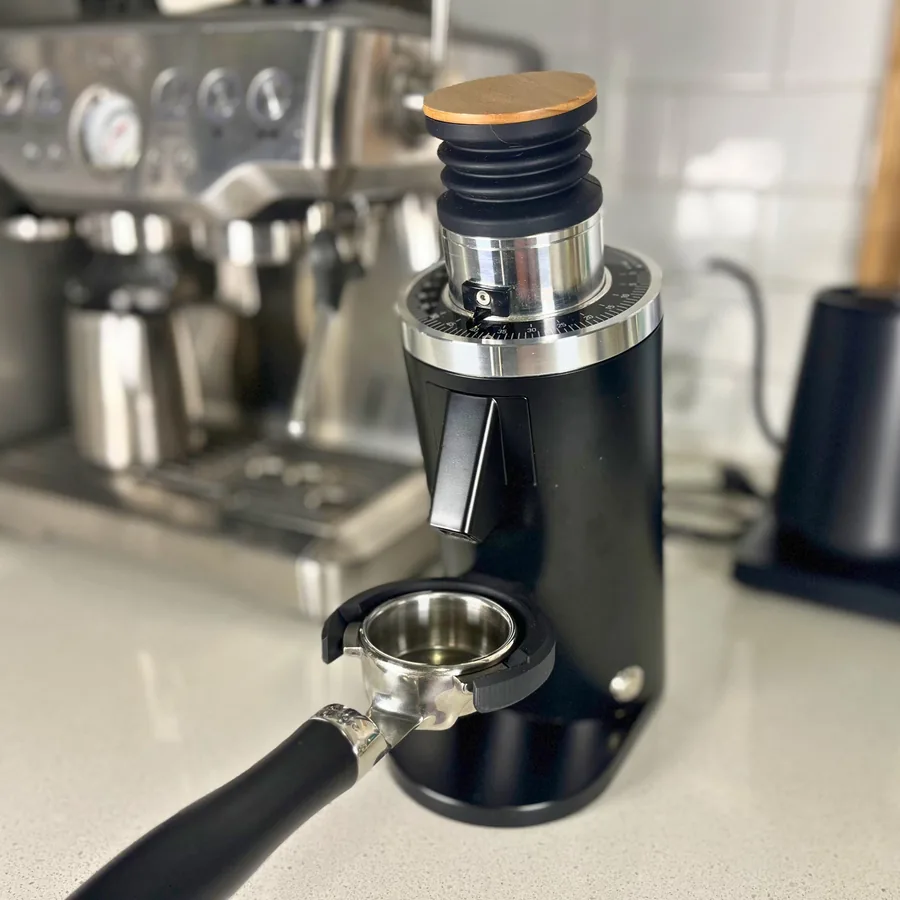 A black 3D-printed fork holding a 54 mm portafilter under the chute of a DF54 grinder, with a Breville espresso machine behind